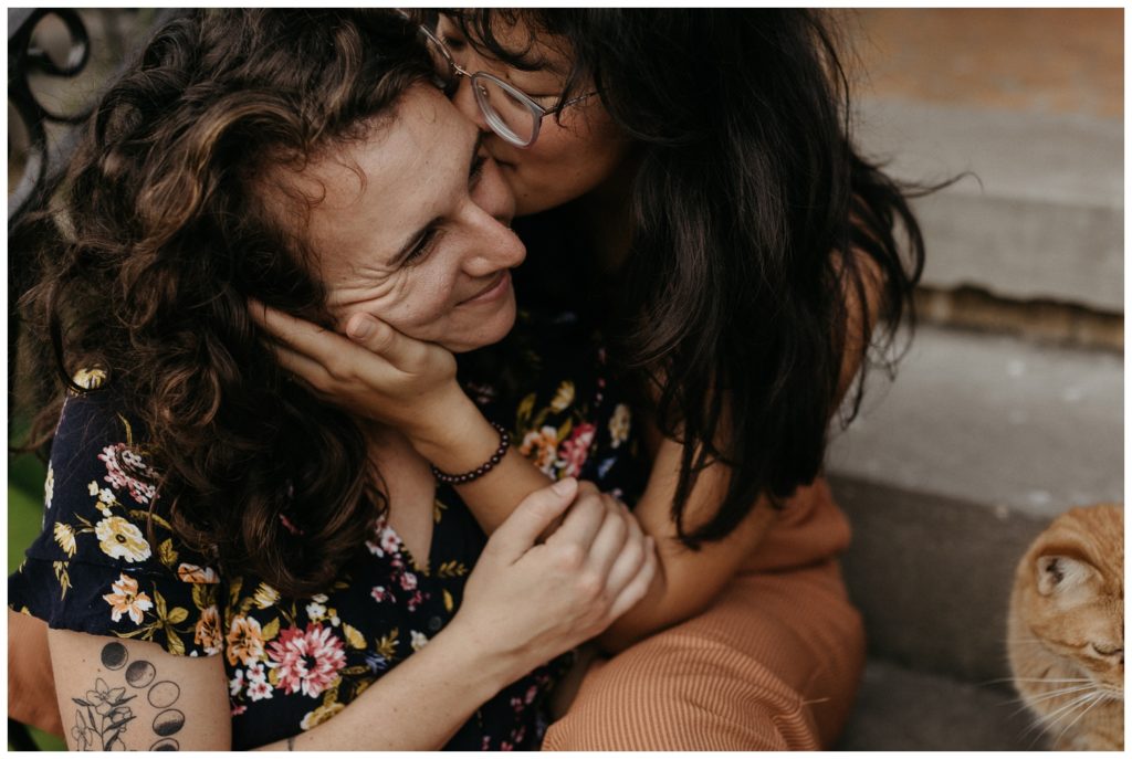 One woman kisses another on the cheek in engagement photos at home