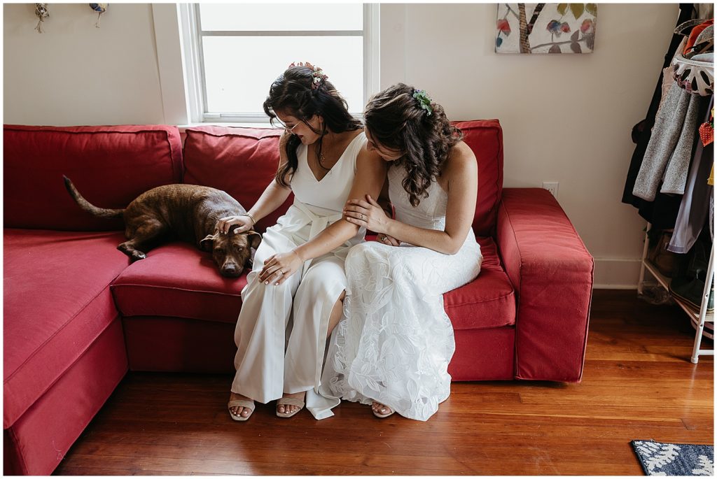 Two women wearing white sit on a red couch petting a dog.