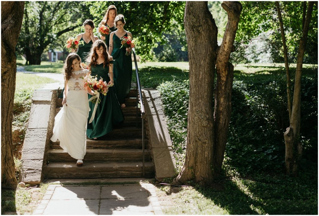 A bride walks down steps with bridesmaids in green dresses.