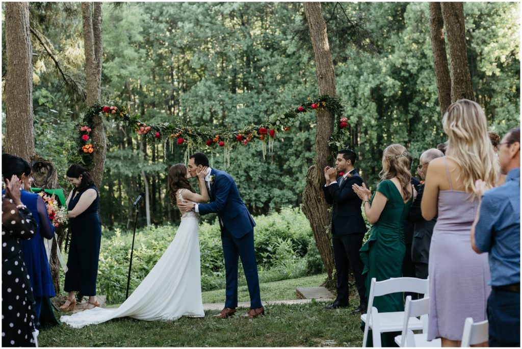 A couple kisses at the end of a wedding ceremony in the woods.