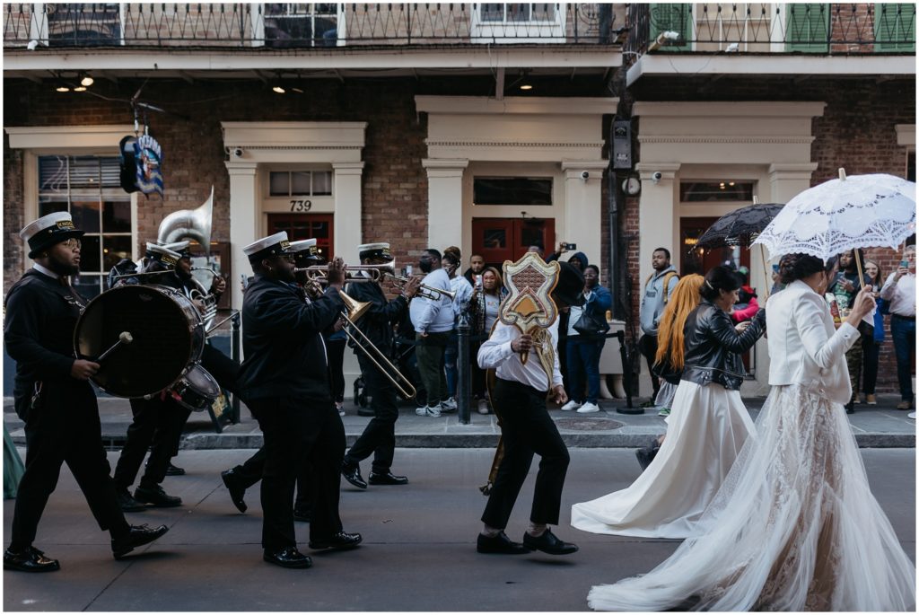 Women in wedding dresses wal  in front of a brass band past a historic building.