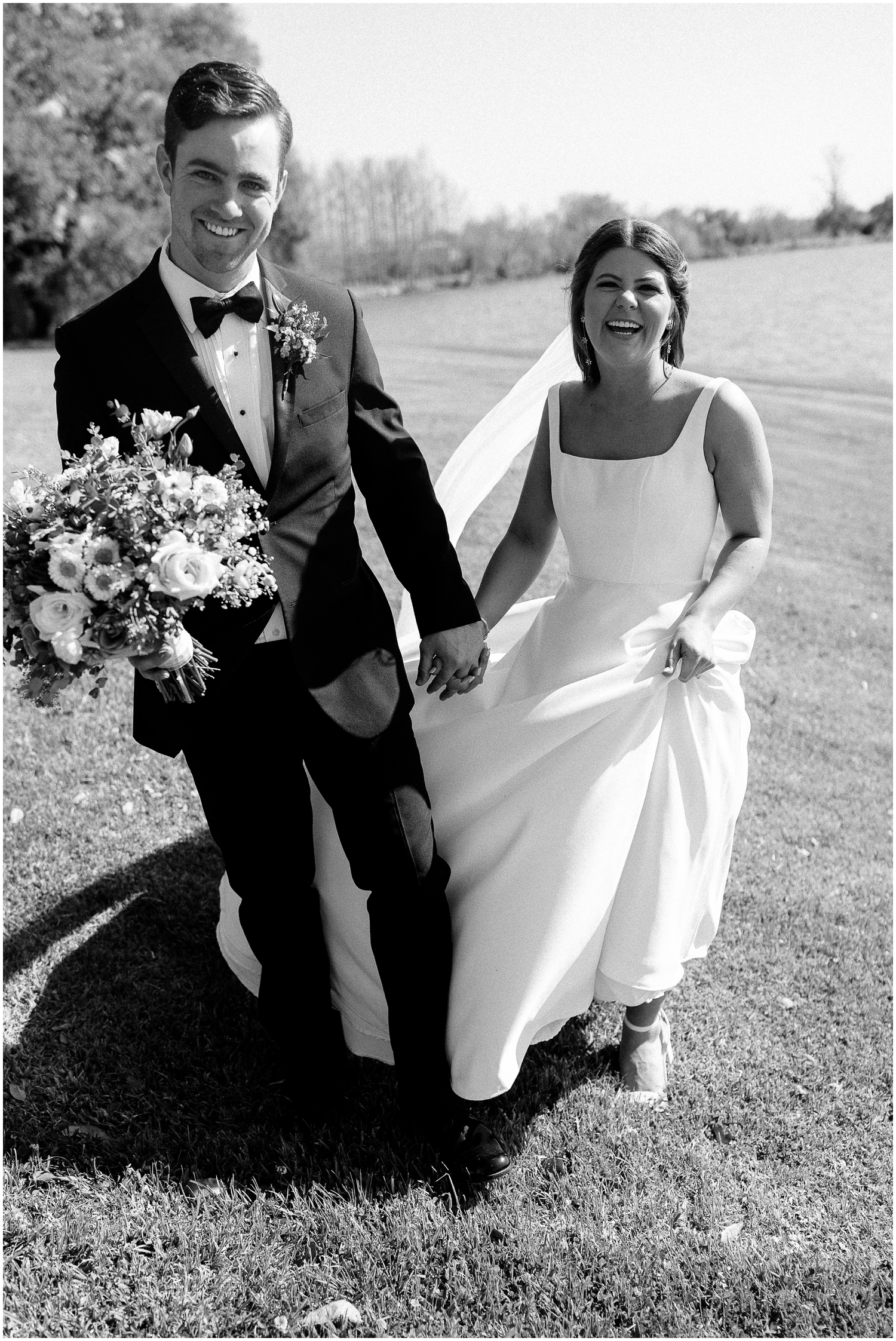A bride and groom hold hands in a field in a black and white wedding photo at Rip Van Winkle Gardens.