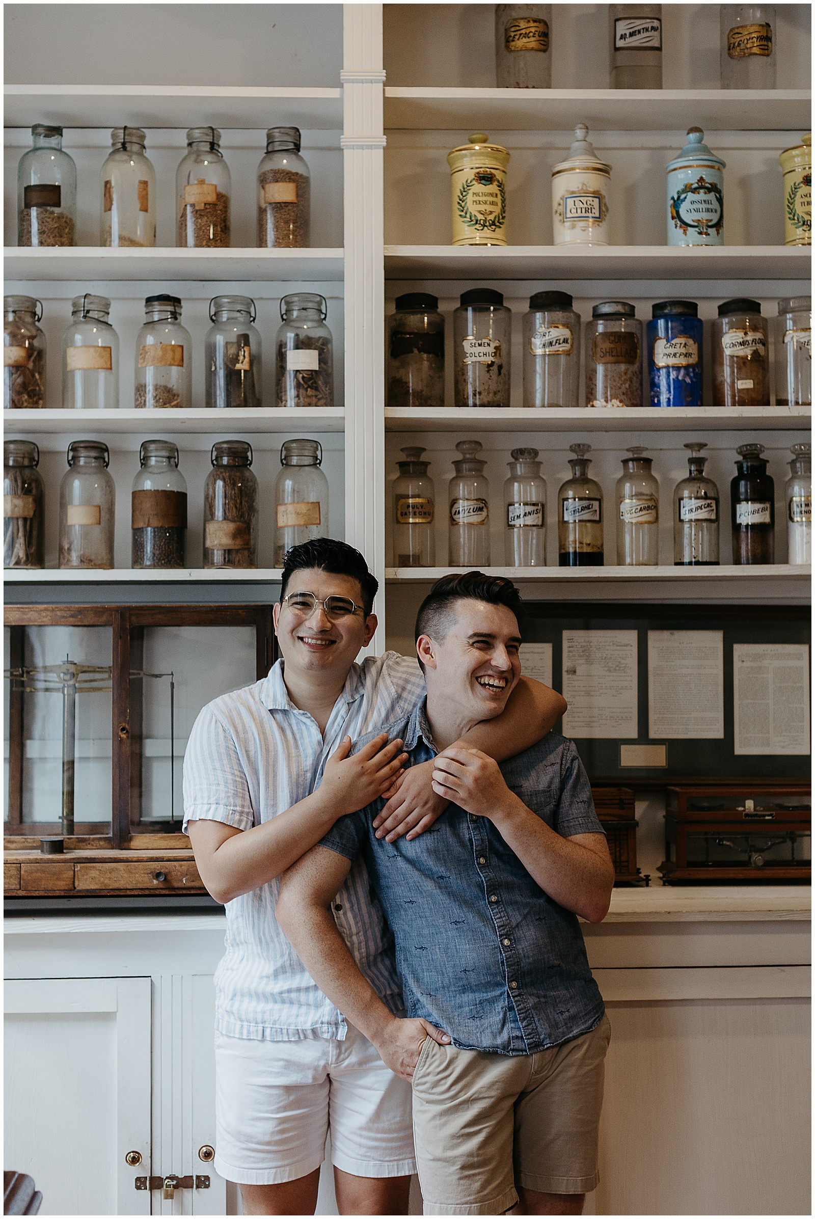 Two men stand in front of a row of Pharmacy Museum shelves for their French Quarter engagement photos.