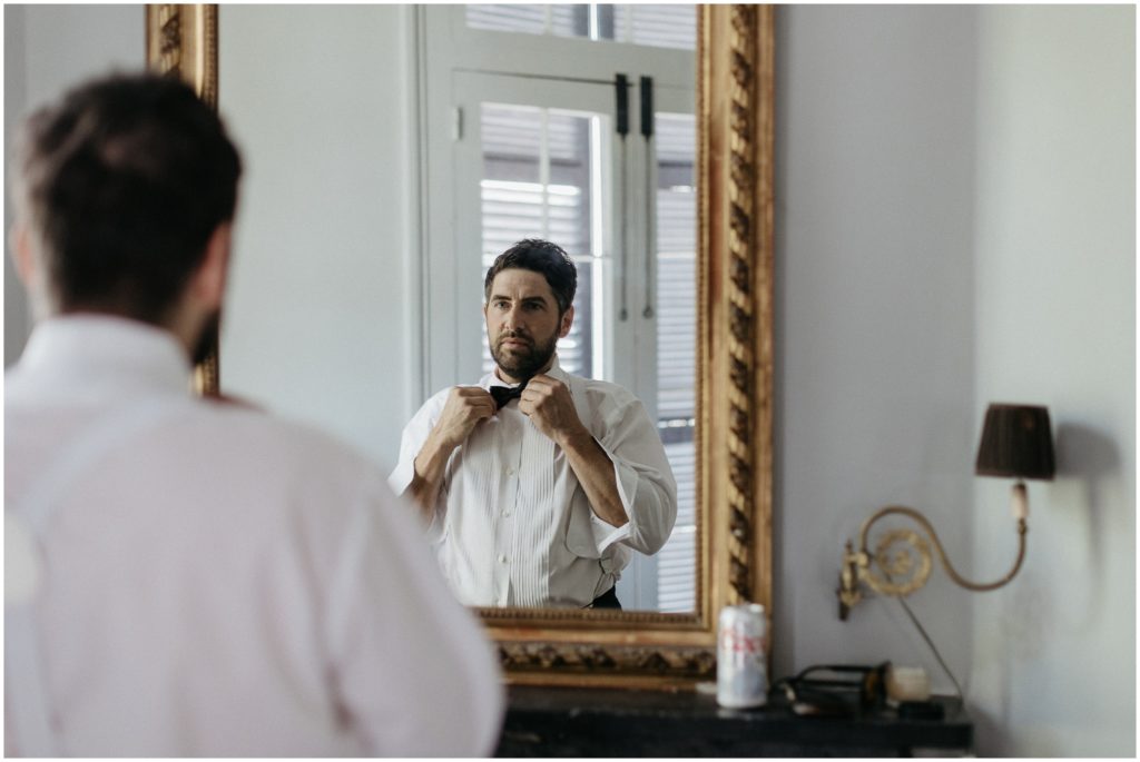 A groom looks in the mirror and straightens his bow tie.