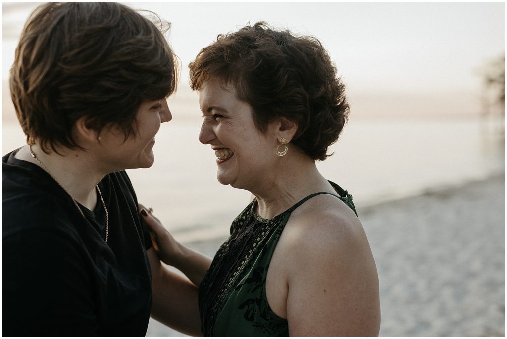 Two people smile at each other on a sandy lake shore.