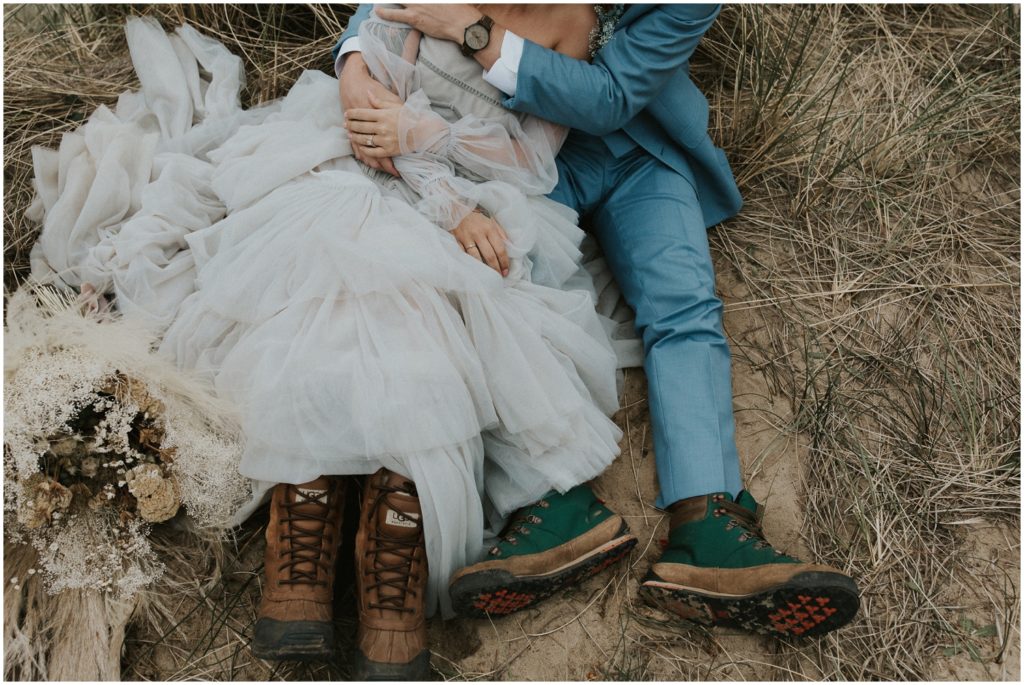 The couple sits close wearing hiking boots with their wedding attire in the sand.