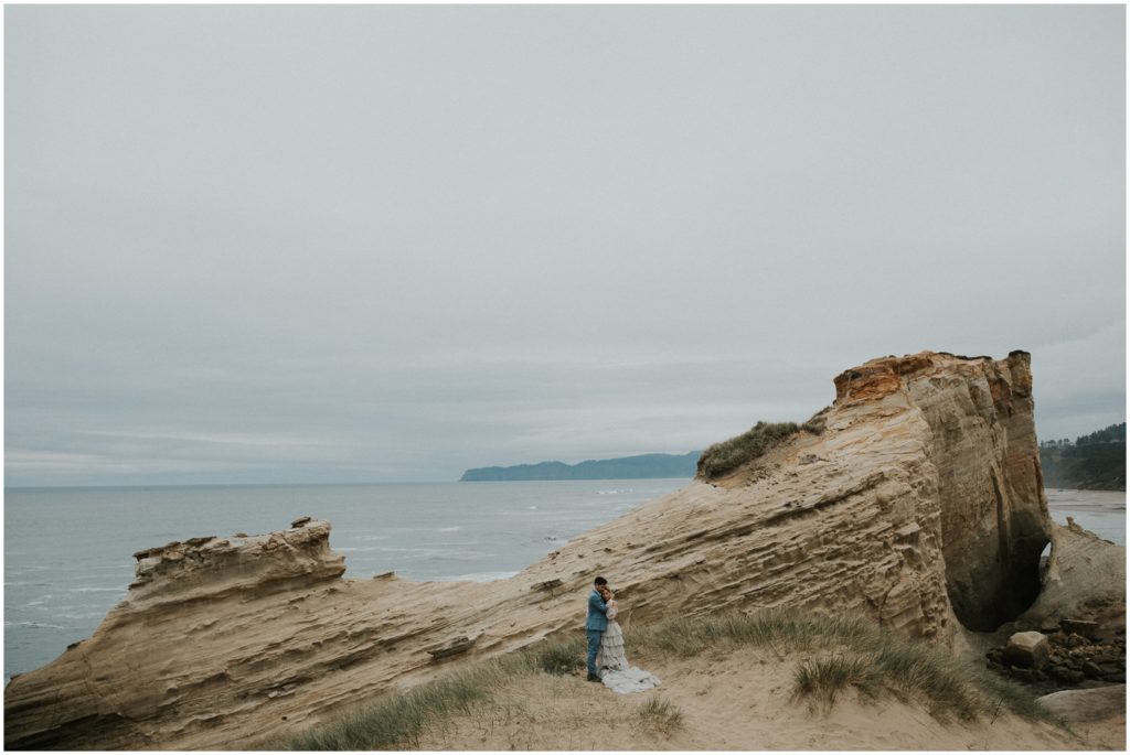 A wedding couple looks over a cliff on a cloudy Oregon beach.