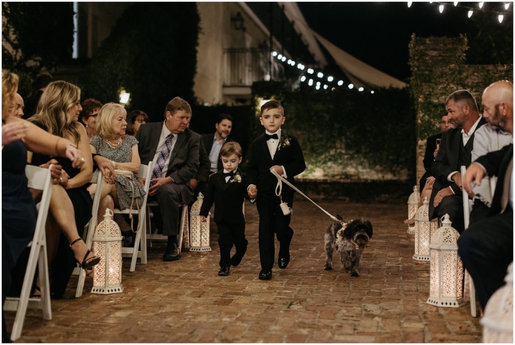 Two boys in suits walk down a wedding aisle guiding a dog on a leash.