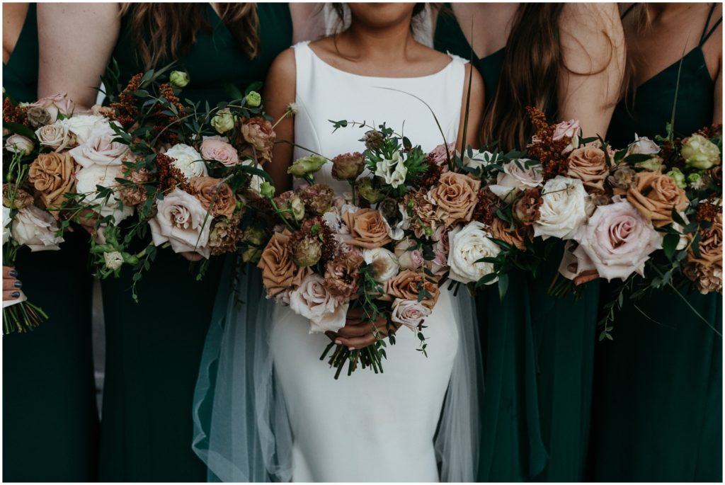A bride and bridesmaids stand in a line holding bouquets of beige and white wedding flowers.