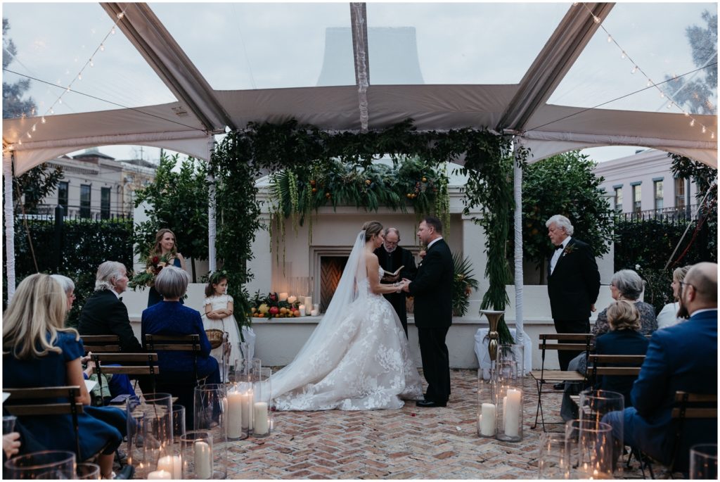 A bride and groom stand beneath an arch of greenery in a courtyard wedding ceremony.