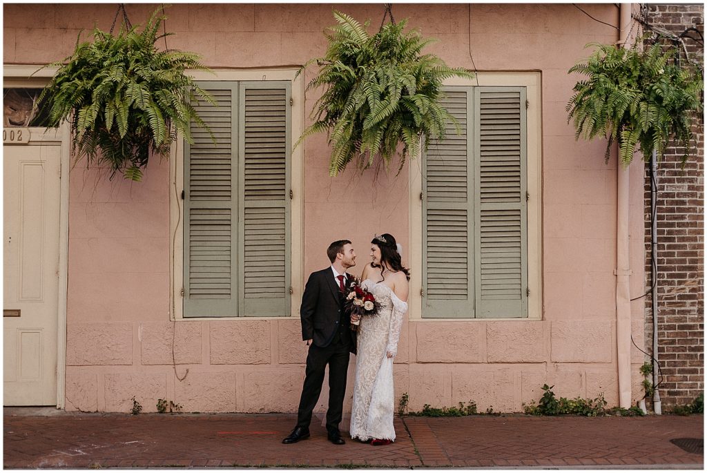 A bride and groom smile at each other in front of a pink French Quarter house.