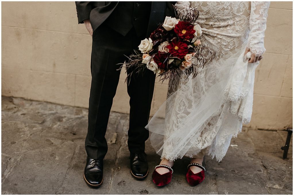 A close up wedding photo shows Hallie's red wedding shoes and gothic wedding bouquet as she stands beside Conor.