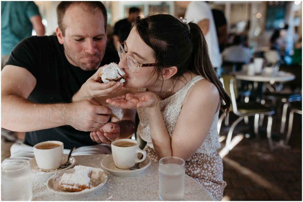 Lauren leans forward over a coffee cup to bite a beignet.