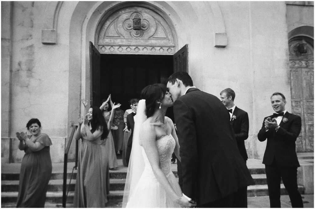 A bride and groom kiss in front of an old church with its doors open in an image taken on disposable cameras for weddings.