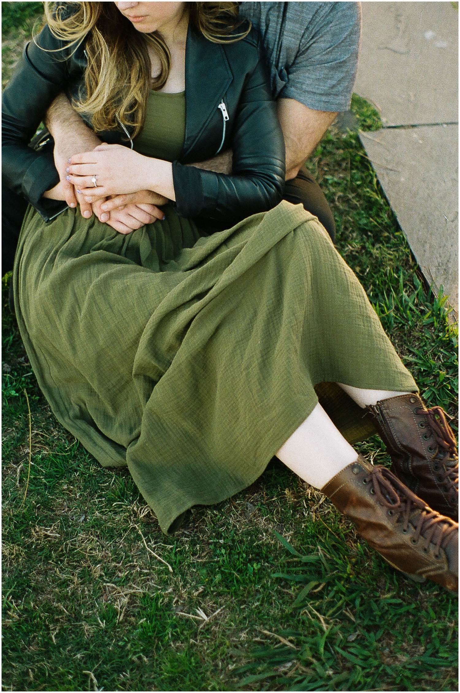 A woman in a green dress and brown boots sits on the grass leaning against a man with her engagement ring showing.