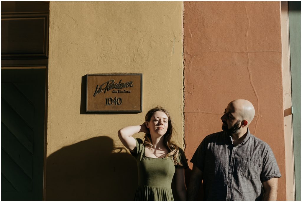 A man and woman stand in front of a brown French Quarter building in a patch of sun for a creative engagement photoshoot.