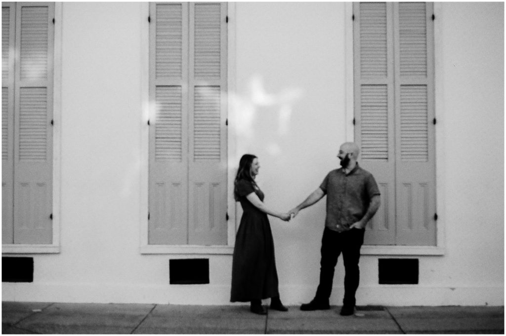 An engaged couple stands in front of a historic New Orleans home holding hands.